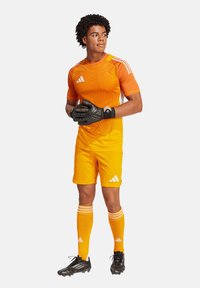 Oranje sporttrui met korte mouwen en diagonale textuurdetails, gecombineerd met bijpassende shorts en kniekousen. Zwarte handschoenen en voetbalschoenen.