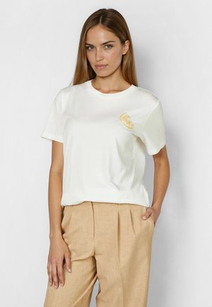 Junge Frau mit langen Haaren, die ein weißes T-Shirt mit kleinem Zitronenmotiv und beige plissierte Hose trägt, steht mit einer Hand in der Tasche.