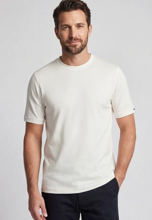 Mann mit Bart, der ein schlichtes weißes T-Shirt mit kurzen Ärmeln und dunkle Hose trägt, steht vor einem hellgrauen Hintergrund.