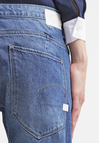 Vaqueros de mezclilla con un tono azul, que presentan un parche de cuero blanco en la cinturilla y dos bolsillos traseros con costuras decorativas.