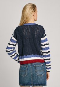 Cardigan tricoté avec des rayures multicolores et un motif en dentelle. Les couleurs principales comprennent le bleu marine, le blanc et le rouge, avec un ourlet côtelé.