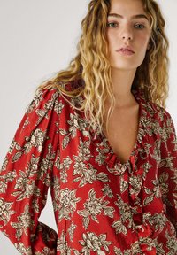 Blouse fleurie rouge avec un col froncé, présentant des motifs de feuilles et de fleurs blancs et beiges. Tissu léger avec des manches amples.