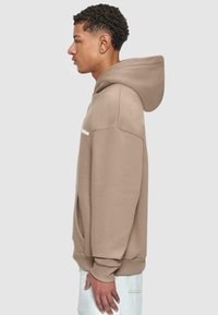Brun hettegenser laget av myk stoff med en romslig hette, dråpe skuldre og frontlommer. Har et minimalistisk hvitt logo på brystet.