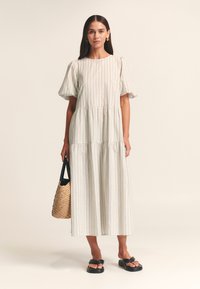 Robe midi gris clair à rayures verticales, manches courtes bouffantes et design à étages. Associée à un sac en paille tressée et des sandales plates noires.