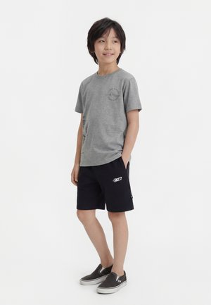 Chlapec stojaci, oblečený v šedej tričku s grafickým motívom, čiernych šortkách s plameniakovým logom a čiernych nazúvacích topánkach, ruky v kapsách, pozerá sa mierne doprava.