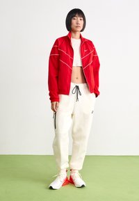 Κόκκινο φερμουάρ μπουφάν με λευκές λεπτομέρειες, λευκό κοντό τοπ, κρεμ φόρμες με τσέπες και λευκά-κόκκινα sneakers σε πρ�άσινο δάπεδο.