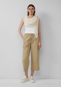 Beige cropped broeken met een elastische tailleband, gecombineerd met een witte aansluitende tanktop en een lichtromige sjaal. De outfit wordt afgemaakt met crèmekleurige sneakers.