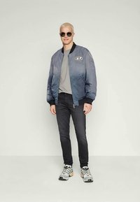 Άνδρας με κοντά ξανθά μαλλιά, φορώντας στρογγυλά γυαλιά ηλίου, μπλε αλεξίπτωτο μπουφάν, γκρι μπλούζα, μαύρα τζιν και γκρι αθλητικά παπούτσια, στέκεται μπροστά σε λευκό φόντο.