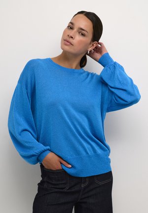 Bright blue, textured knit sweater with a round neckline, dropped shoulders, and gathered cuffs. Paired with dark jeans.