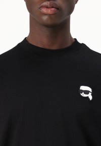 Tricou negru cu mânecă scurtă, având un pin alb în stil cartoon al unei persoane cu ochelari de soare în zona stânga a pieptului. Textură moale din bumbac.