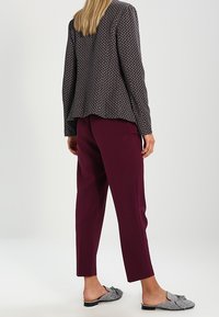 Kvinna i mönstrad kavaj, plommonfärgade ankel-långa byxor och grå rutiga slip-on skor med rosetter, som står framför en enfärgad bakgrund.