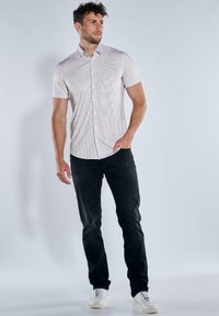 Herren Kurzarmhemd in hellem Beige mit dezenten Streifen, kombiniert mit schwarzen Slim-Fit-Jeans und weißen Sneakers. Glatter Stoff, Button-Down-Kragen.