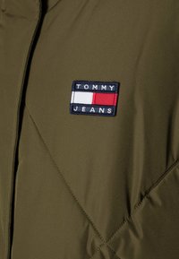 Olive green puffer jacket featuring a quilted texture and a rectangular blue and red "TOMMY JEANS" logo patch on the chest.