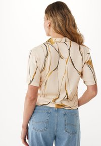 Korte mouwen blouse in lichtbeige met een marmerpatroon in goud en zwart. Beschikt over een kraag en een gladde, lichte stof.