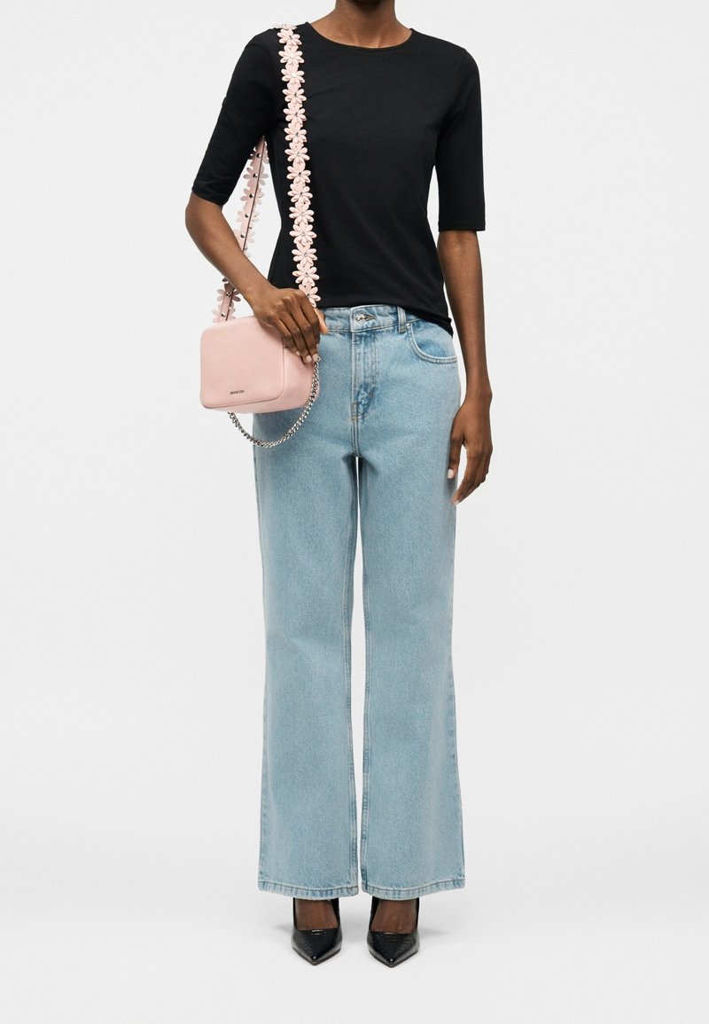 Personne portant un top noir, un jean bleu clair à jambes larges, des chaussures noires pointues, et tenant un sac à bandoulière rose avec une sangle fleurie.