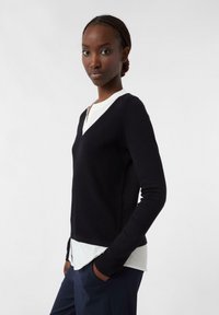 Junge Frau mit geflochtenem Haar, die einen schwarzen V-Ausschnitt-Pullover über einer weißen Bluse und einer marineblauen Hose trägt, mit den Händen in den Taschen stehend.