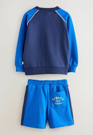 Sinine spordikomplekt, mis sisaldab pulloveri särk, millel on tumesinised ja helesinised aktsendid, ning vastavaid šortse, millel on "SPORTS 1974 LEAGUE" graafika taskul.