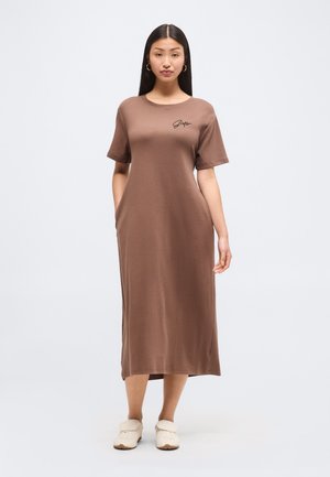 Kobieta stoi, ubrana w brązową sukienkę midi z krótkimi rękawami, czarne logo na piersi, białe buty wsuwane i złote kolczyki koła.