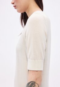 Jasnobeżowy sweter z krótkim rękawem, okrągłym dekoltem i prążkowaniem na mankietach. Tkanina wydaje się miękka, z widoczną fakturą i szwami.