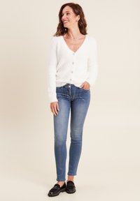 Cardigan bianco fluffy con chiusura a bottoni, abbinato a jeans skinny blu. Mocassini neri completano l'outfit. Design semplice, texture accogliente.