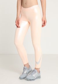 Różowe legginsy Nike w kolorze brzoskwiniowym z błyszczącą fakturą, z wysokim pasem i srebrnym znakiem Swoosh akcentującym bok, połączone z szarymi butami sportowymi.
