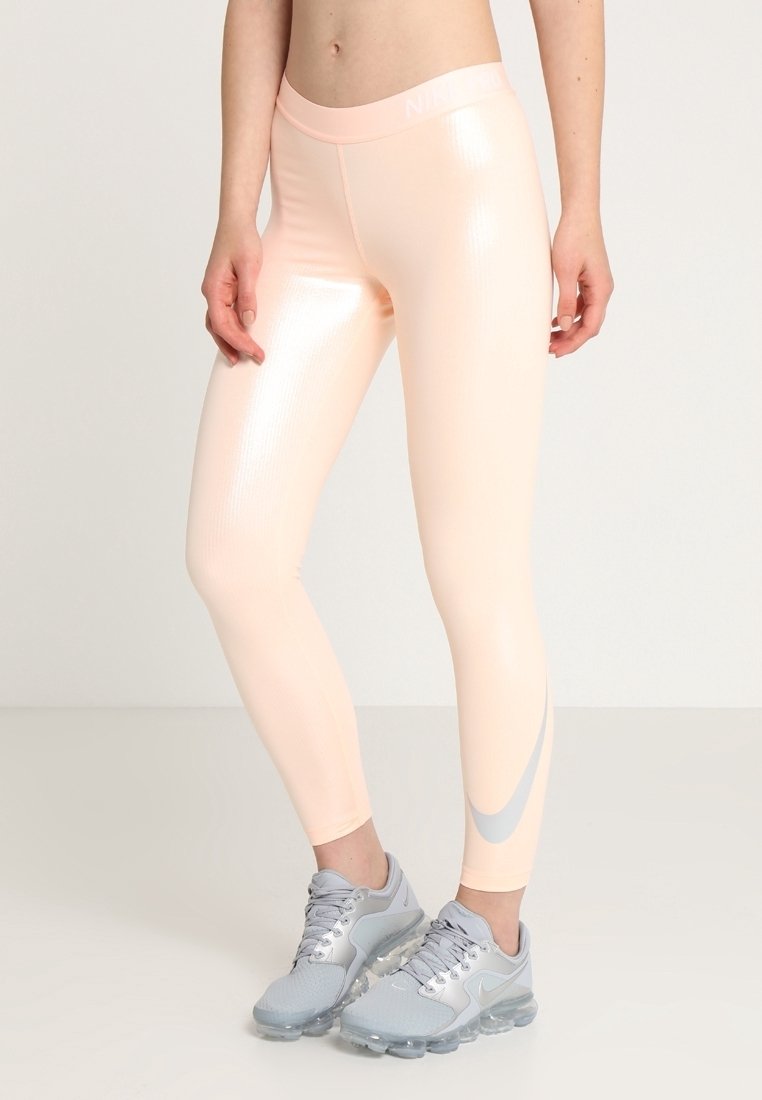 Różowe legginsy Nike w kolorze brzoskwiniowym z błyszczącą fakturą, z wysokim pasem i srebrnym znakiem Swoosh akcentującym bok, połączone z szarymi butami sportowymi.