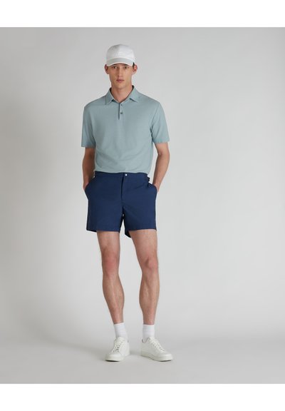 Hellblaues Polohemd mit Kragen, marineblaue Shorts und weiße Sneaker. Das Model trägt eine weiße Kappe und steht mit Händen in den Taschen.