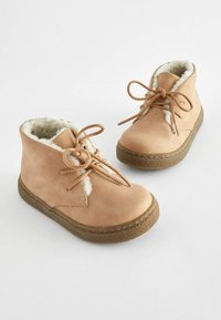 Bottines en daim beige avec une doublure en fourrure douce, bout rond et semelles en caoutchouc. Présente un système de laçage et des accents de couture visibles.