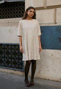 Bēša, fakturēta kleita ar īsām piedurknēm, ceļa garumā, brīva piegriezuma. Valkāta ar melnām necaurspīdīgām zeķbiksēm un tumšām kurpēm.