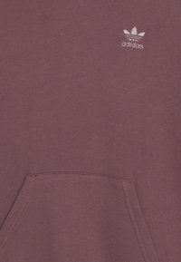 Bordová mikina s měkkou texturou, která má klokaní kapsu a malou bílou značku Adidas v levém horním rohu.