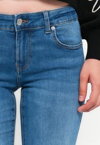 Jeans de denim azul ajustados con cintura media, sutiles desgastes y costuras en contraste. Presenta bolsillos frontales y cierre de botón metálico.
