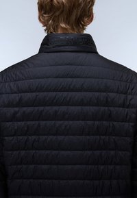 Personne aux cheveux courts châtain clair portant une veste matelassée noire, montrant le dos et les détails du col.