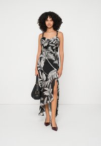 Kvinna med lockigt hår, iförande en svart och vit blommig klänning med en slits, bärande en svart handväska och burgundyröda klackar, stående mot vit bakgrund.