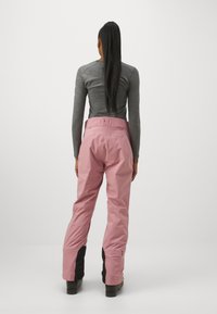 Pantalons de ski imperméables roses avec une taille haute, en tissu texturé, et une finition noire au niveau des chevilles. Vu de l'arrière, présentant un design lisse.
