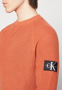 Orange stickad tröja med rund halsringning, ribbad textur och svart Calvin Klein Jeans-logotyppatch på vänster ärm.