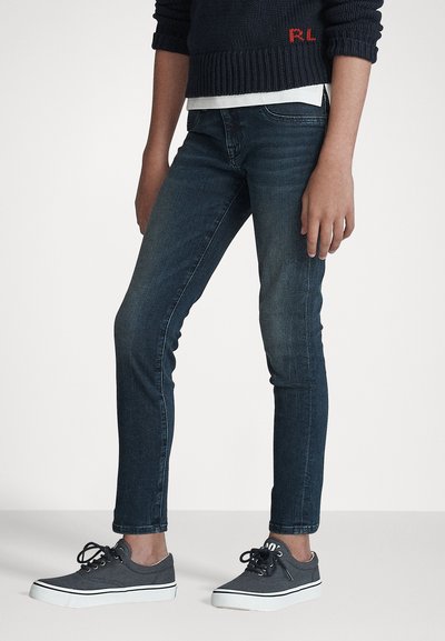 Jeans ajustados de color azul oscuro con una textura suave y un ligero desvanecimiento. Combinados con zapatillas de lona grises que tienen suelas blancas y cordones negros.