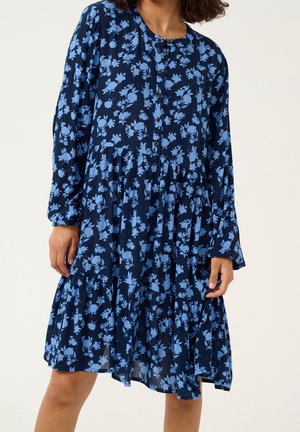 Donna che indossa un vestito a maniche lunghe blu navy con stampa floreale azzurro chiaro e gonna a balze, in piedi davanti a uno sfondo uniforme.