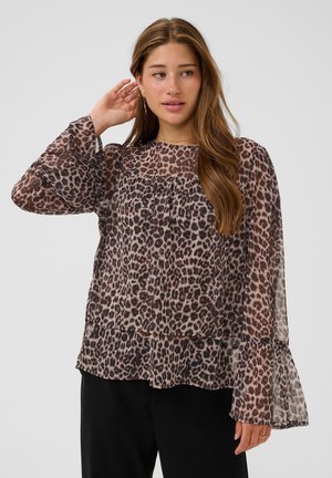 Jonge vrouw met lang bruin haar draagt een doorschijnende blouse met lange mouwen en luipaardprint en zwarte broek, raakt een oor aan en kijkt opzij.
