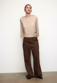Bež širok pulover z veliko čolnasto izrezom, kombiniran s rjavimi kavbojkami z ravnimi hlačnicami. Gladek tekstura tkanine, minimalističen dizajn, brez vidnih vzorcev.