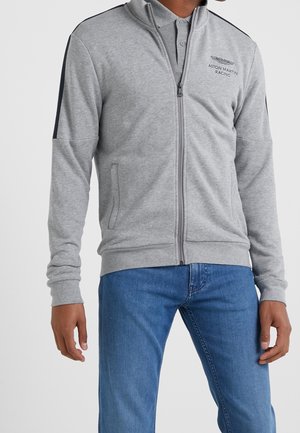 Mężczyzna w jasnoszarej kurtce na zamek Aston Martin Racing z czarnym paskiem na rękawach i niebieskich dżinsach, stojący na jednolitym tle.