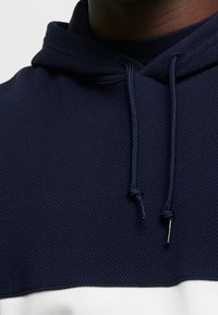 Tmavě modrá mikina s kapucí z texturované tkaniny, nastavitelnými šňůrami a bílým vodorovným pruhem podél dolního okraje.