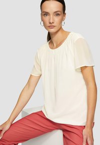 Weißes Oberteil mit kurzen, transparenten Ärmeln, rundem Halsausschnitt und Raffung; kombiniert mit korallfarbenen Hosen vor einem schlichten Hintergrund.