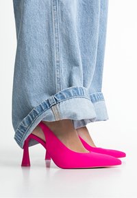 Różowe buty na obcasie typu slingback o gładkim wykończeniu i spiczastym nosku, noszone z jasnoniebieskimi, podwiniętymi dżinsami. Krój z rozszerzanym obcasem.