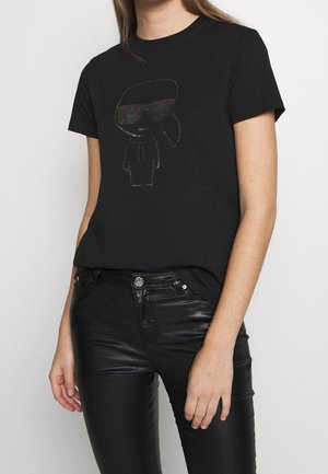 Musta t-paita, jossa on graafinen kuvio hahmosta, jolla on suurennetut aurinkolasit. Valmistettu puuvillasta, rento istuvuus, yhdistetty mustiin kapealahkeisiin housuihin.