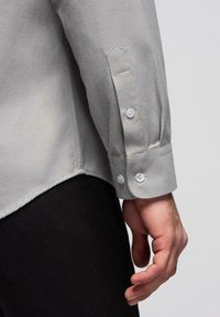 Bras gauche d'une personne portant une chemise à manches longues gris clair avec des poignets boutonnés et un pantalon noir, sur un fond uni.