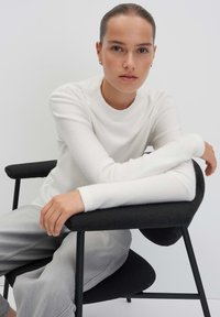 Weißes, geripptes Langarmshirt mit rundem Ausschnitt, kombiniert mit hellgrauen Hosen, sitzt auf einem schwarzen gepolsterten Stuhl. Glatte Textur, minimalistisches Design.