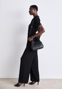 Kobieta ubrana w czarny kombinezon i szpilki, niosąca czarną skórzaną torebkę z złotym logo na pasku na ramię.