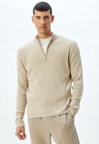 Suéter beige de canalé con un collar de media cremallera, hecho de material suave. Mangas largas y un corte relajado, combinado con pantalones beige claros.