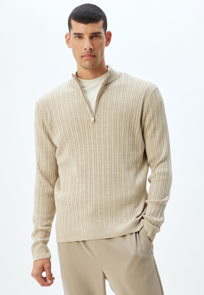 Suéter beige de canalé con un collar de media cremallera, hecho de material suave. Mangas largas y un corte relajado, combinado con pantalones beige claros.