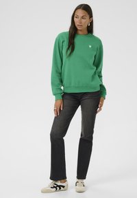 Grüner Pullover mit einem kleinen Herz-Logo auf der Brust, kombiniert mit dunklen Jeans und cremefarbenen Sneakers mit schwarzen Streifen.